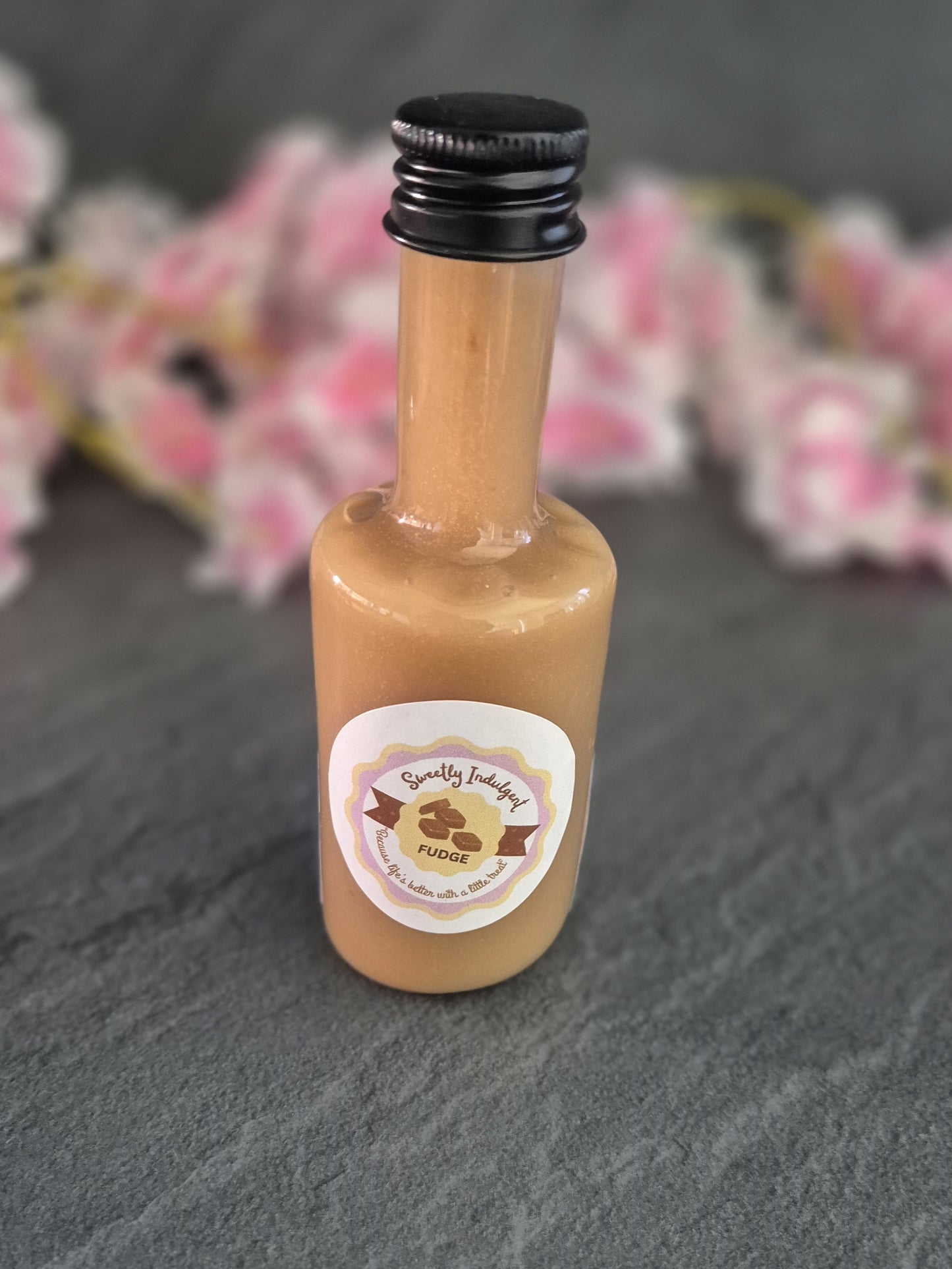 Small fudge sauce bottle with a label on a dark surface with blurred pink flowers in the background