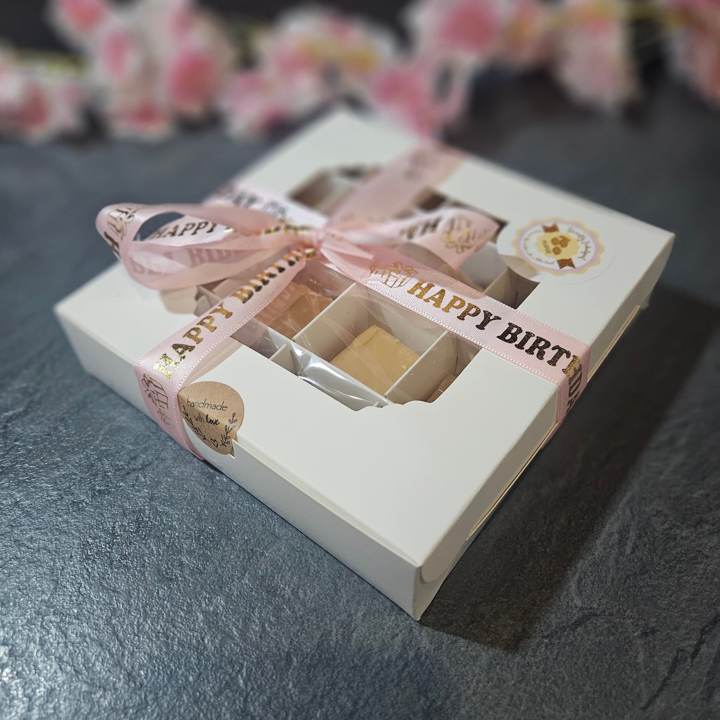 Gift box with Happy Birthday pink ribbon on a dark surface with blurred floral background