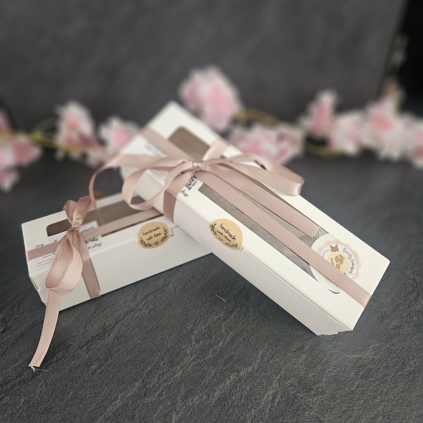 Two white gift boxes with pink ribbons and handmade with love labels on a dark surface with blurred floral background