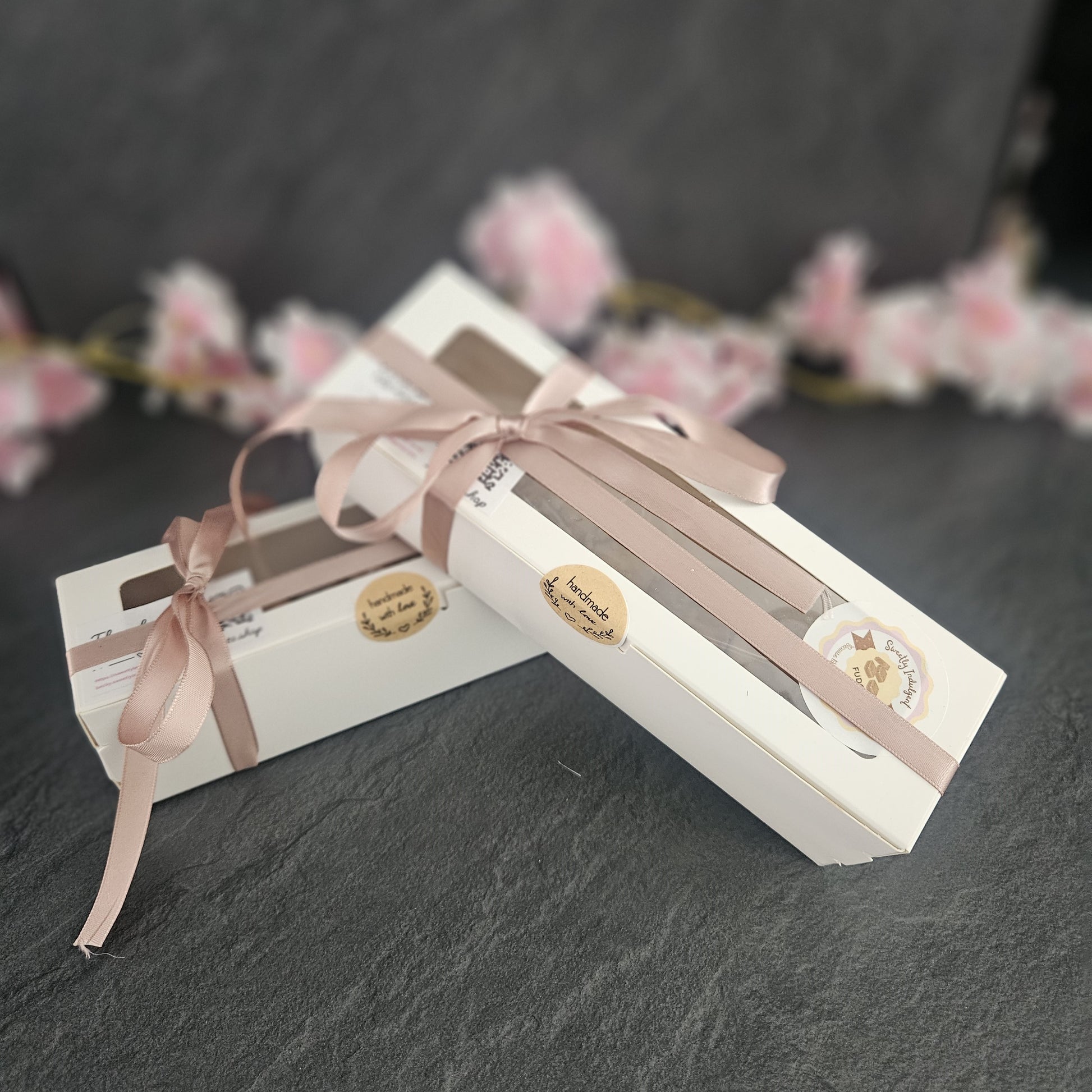 Two white gift boxes with pink ribbons and handmade with love labels on a dark surface with blurred floral background