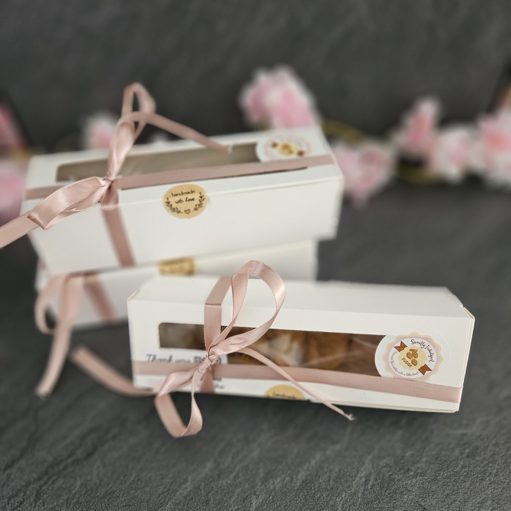Three white gift boxes with pink ribbons on a dark surface sealed with brown sticker and cherry blossoms in background.