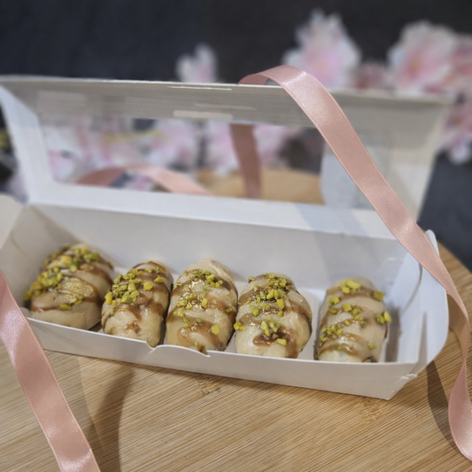 Open white rectangular box containing 5 fudge dipped dates with a pink ribbon on a wooden surface.