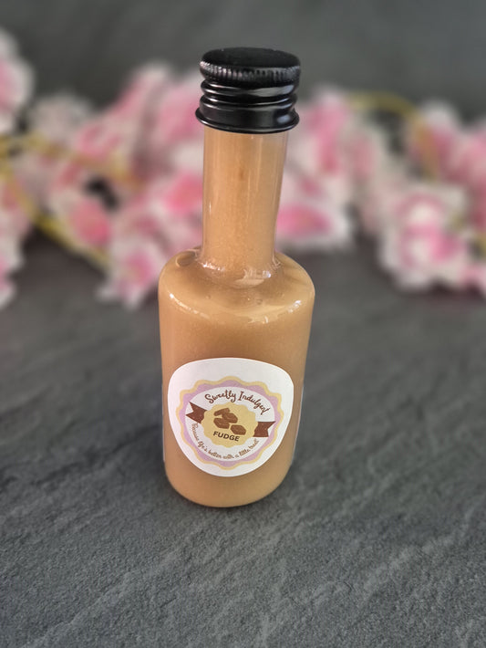 Small fudge sauce bottle with a label on a dark surface with blurred pink flowers in the background