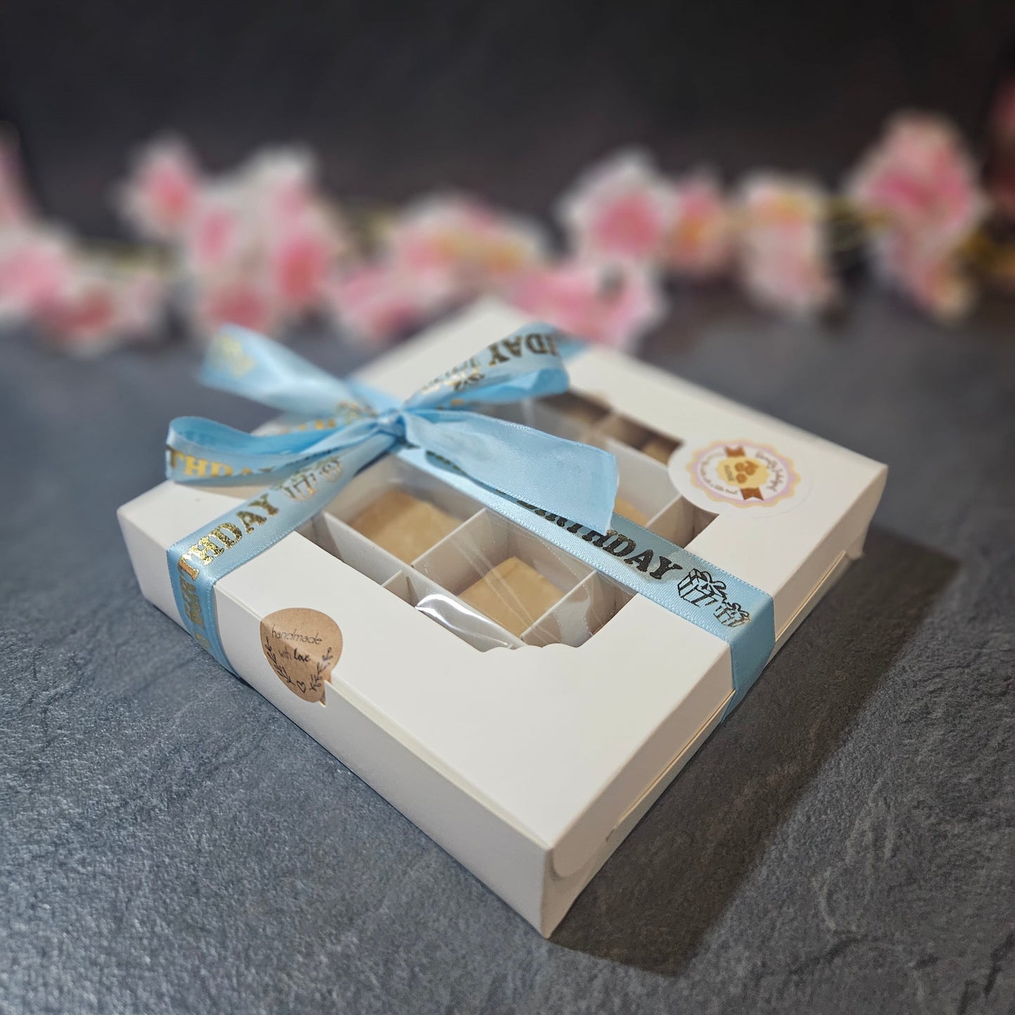 Gift box with a blue ribbon and Happy Birthday text on a dark surface with blurred flowers in the background.