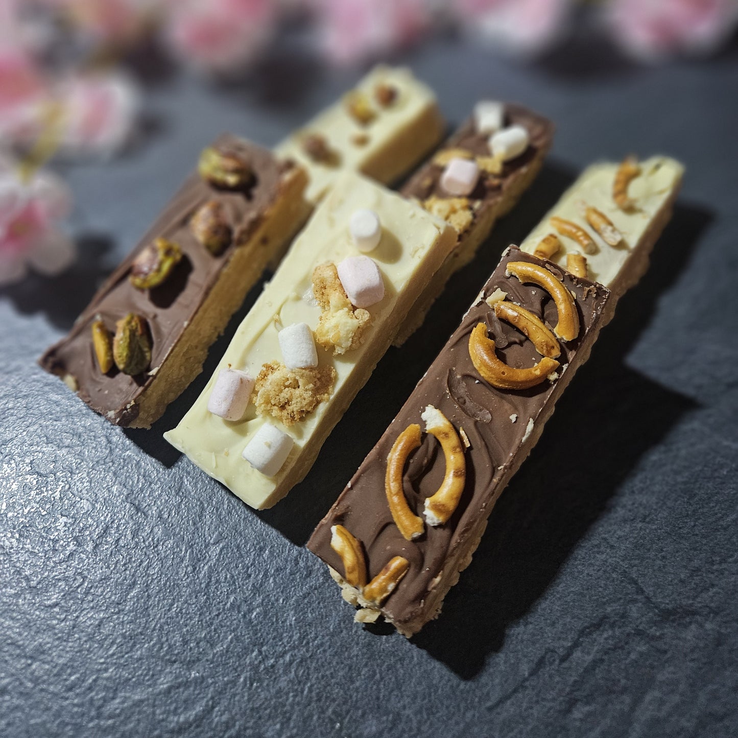 Three chocolate-dipped fudge bars with various toppings on a dark surface. with blurred flowers in background