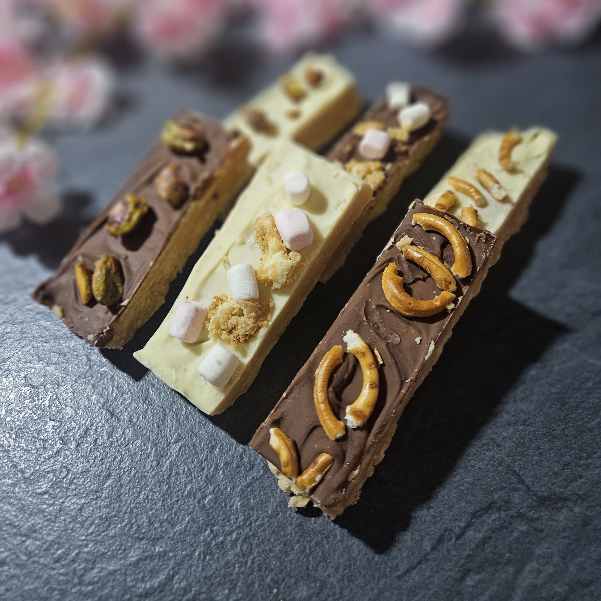 Three chocolate-dipped fudge bars with various toppings on a dark surface. with blurred flowers in background