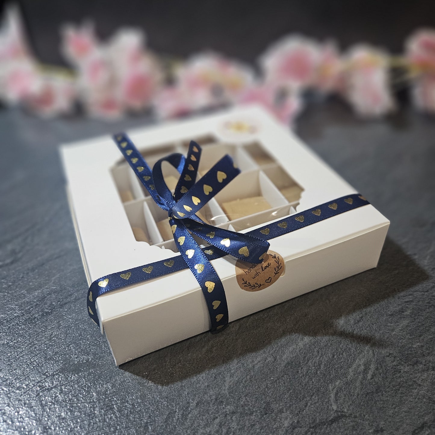 White gift box with a blue ribbon on a dark surface with blurred flowers in the background. sealed with handmade with love sticker.