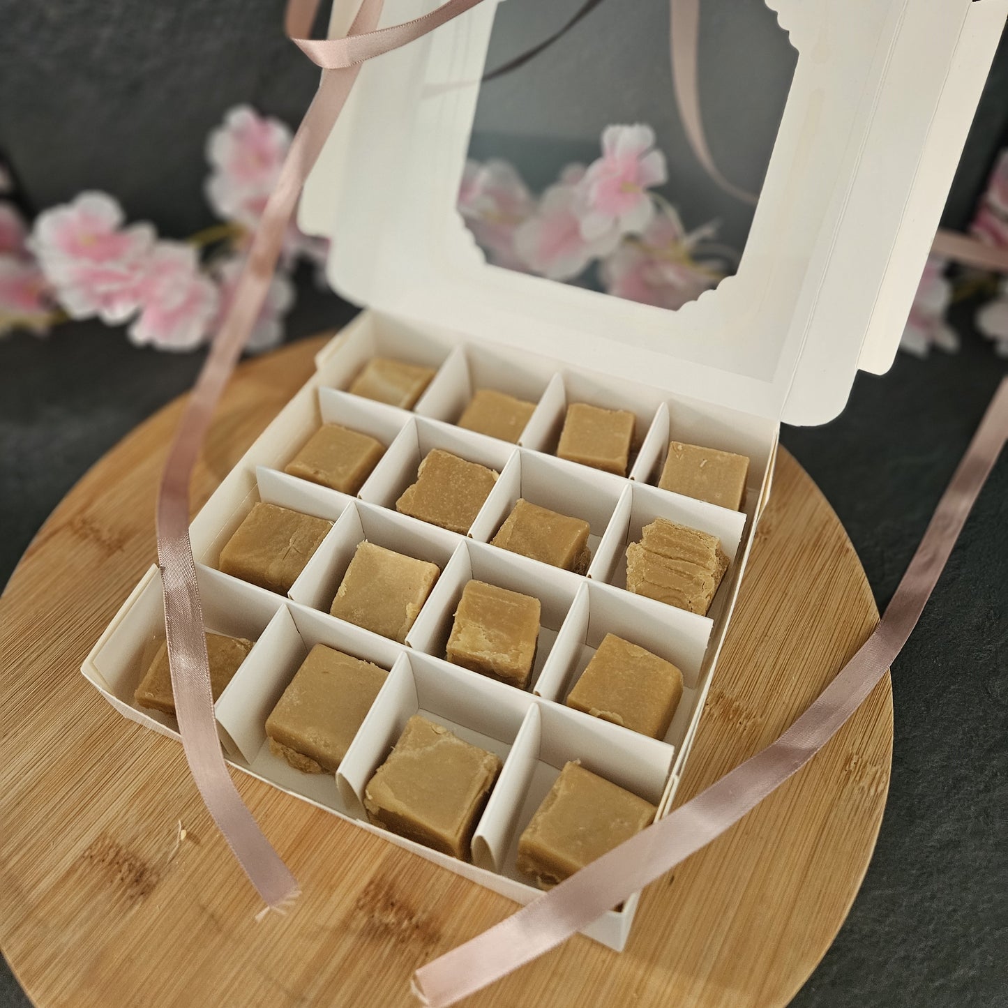 Large box of traditional fudge with a pink ribbon on a wooden surface and flowers in the background.