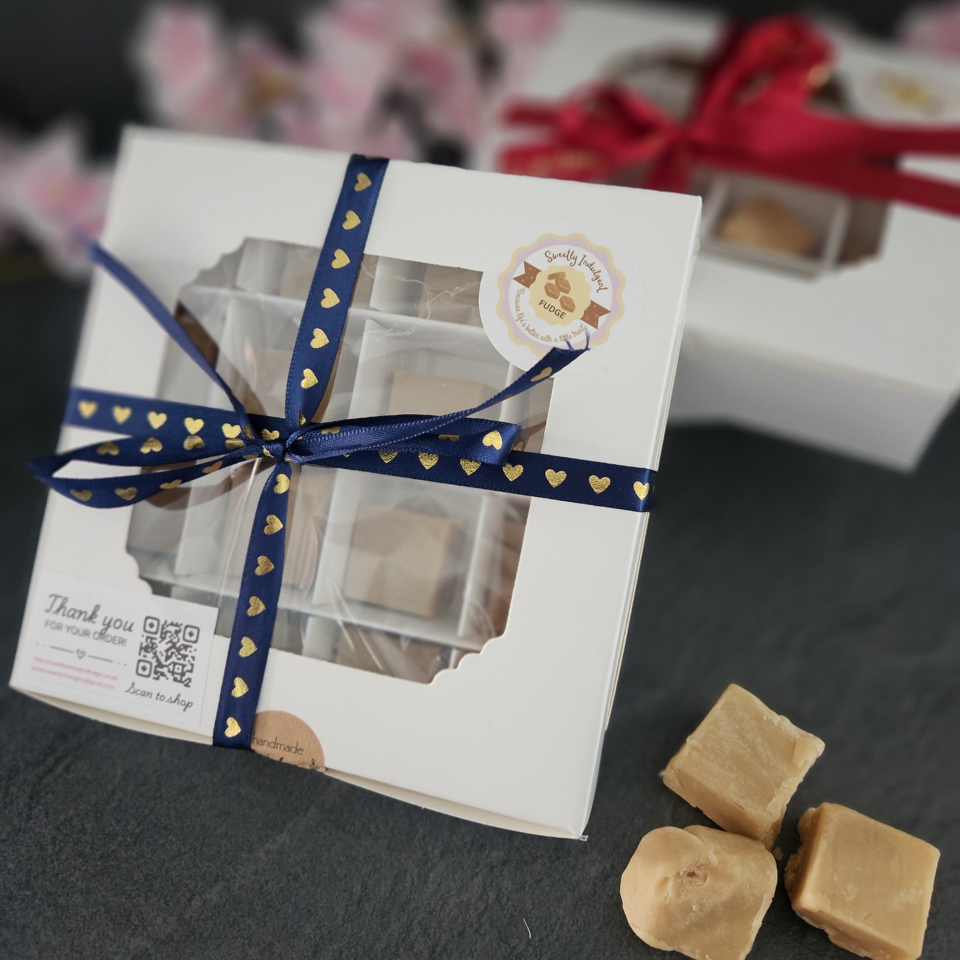 Gift box with a blue ribbon with gold hearts, accompanied by some fudge on a dark surface. second box in background