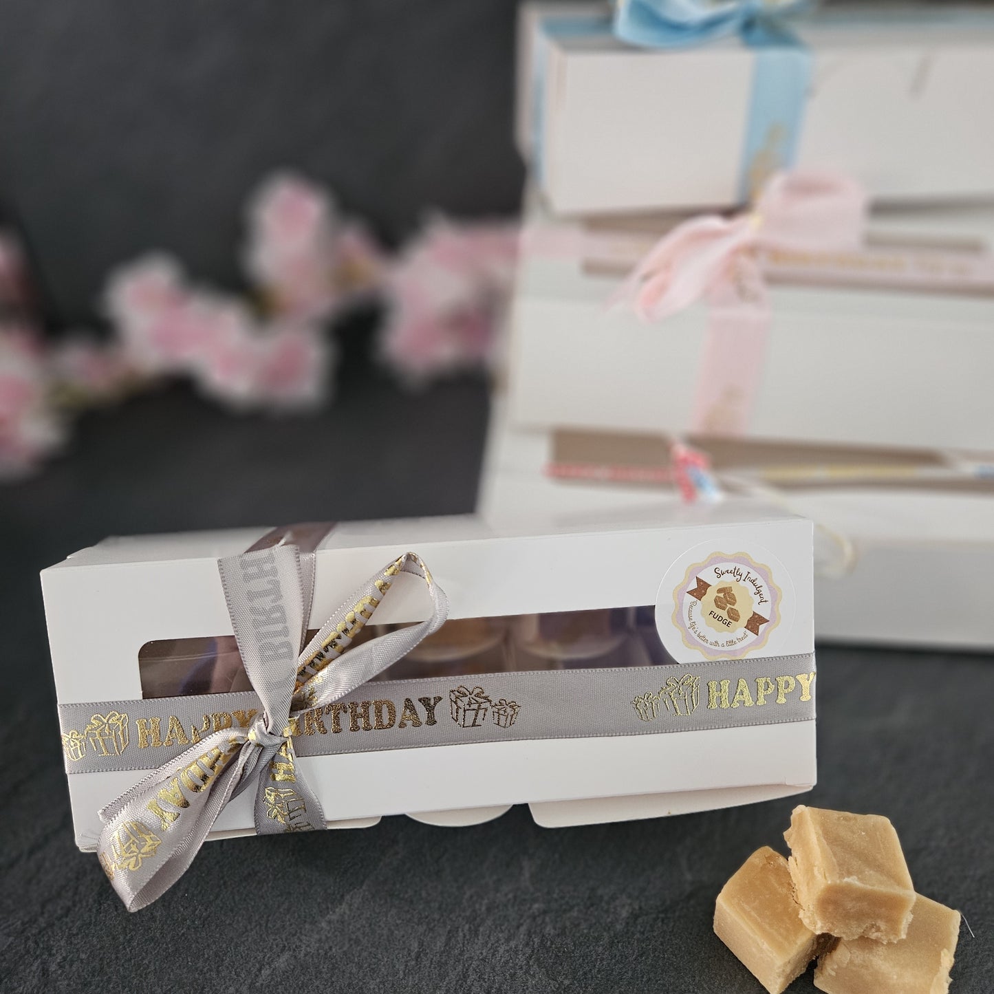 Box of fudge with a ribbon on a dark surface, with blurred gift boxes and flowers in the background.
