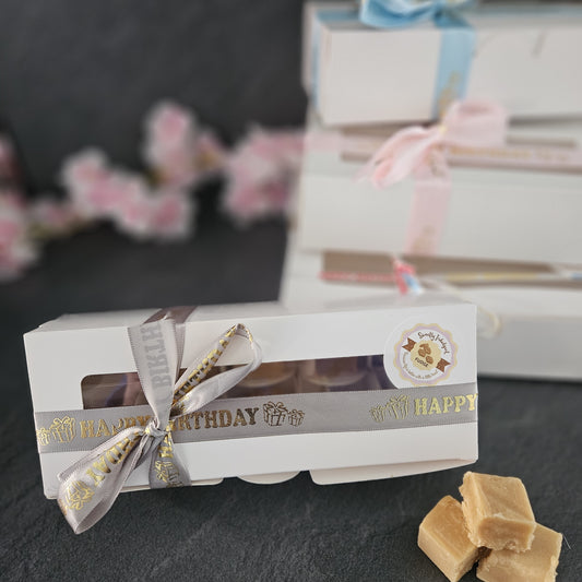Box of fudge with a ribbon on a dark surface, with blurred gift boxes and flowers in the background.