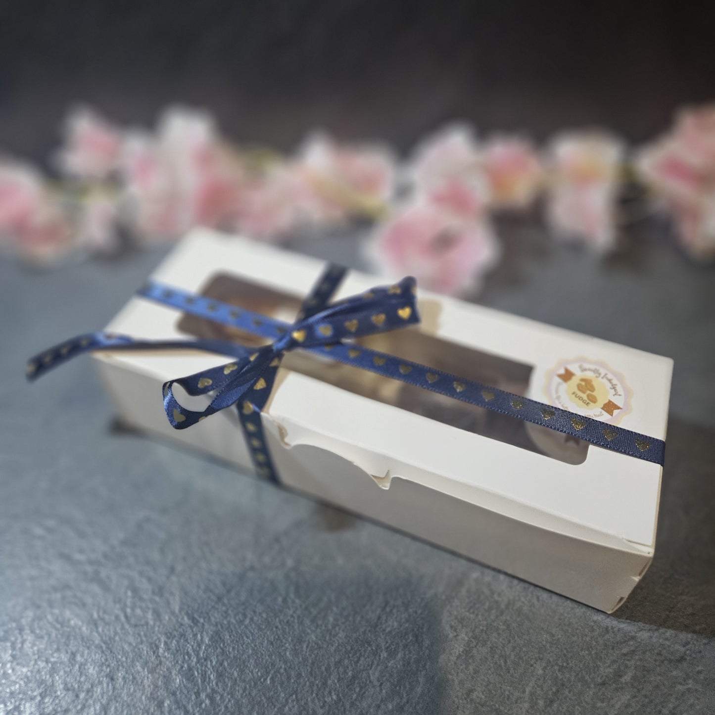 Gift box with a blue ribbon on a dark surface with blurred flowers in the background