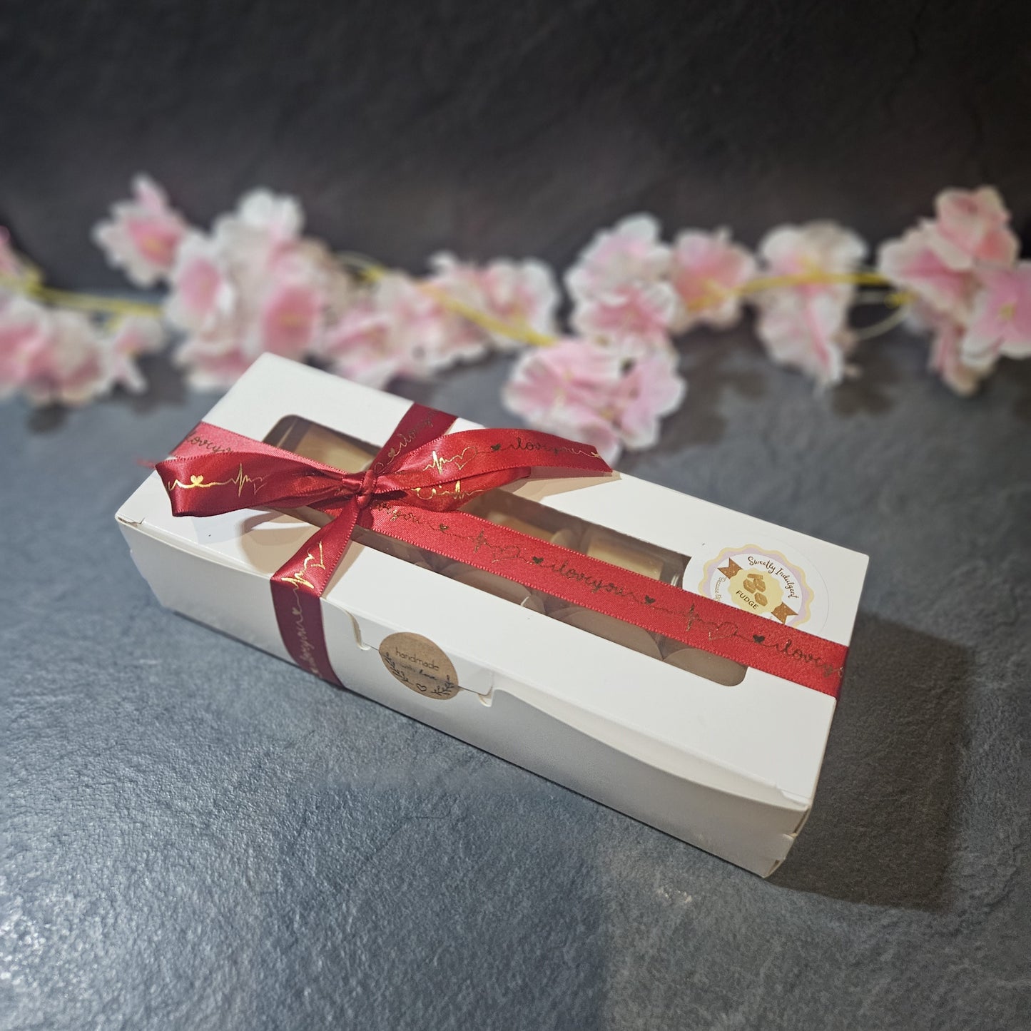 Gift box with a red ribbon saying I love you on a dark surface with pink flowers in the background. sealed with made with love sticker.