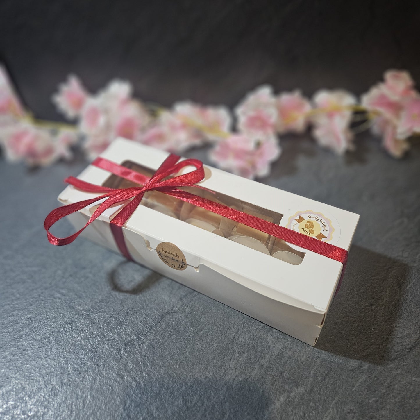Gift box with a red ribbon on a gray surface with blurred flowers in the background