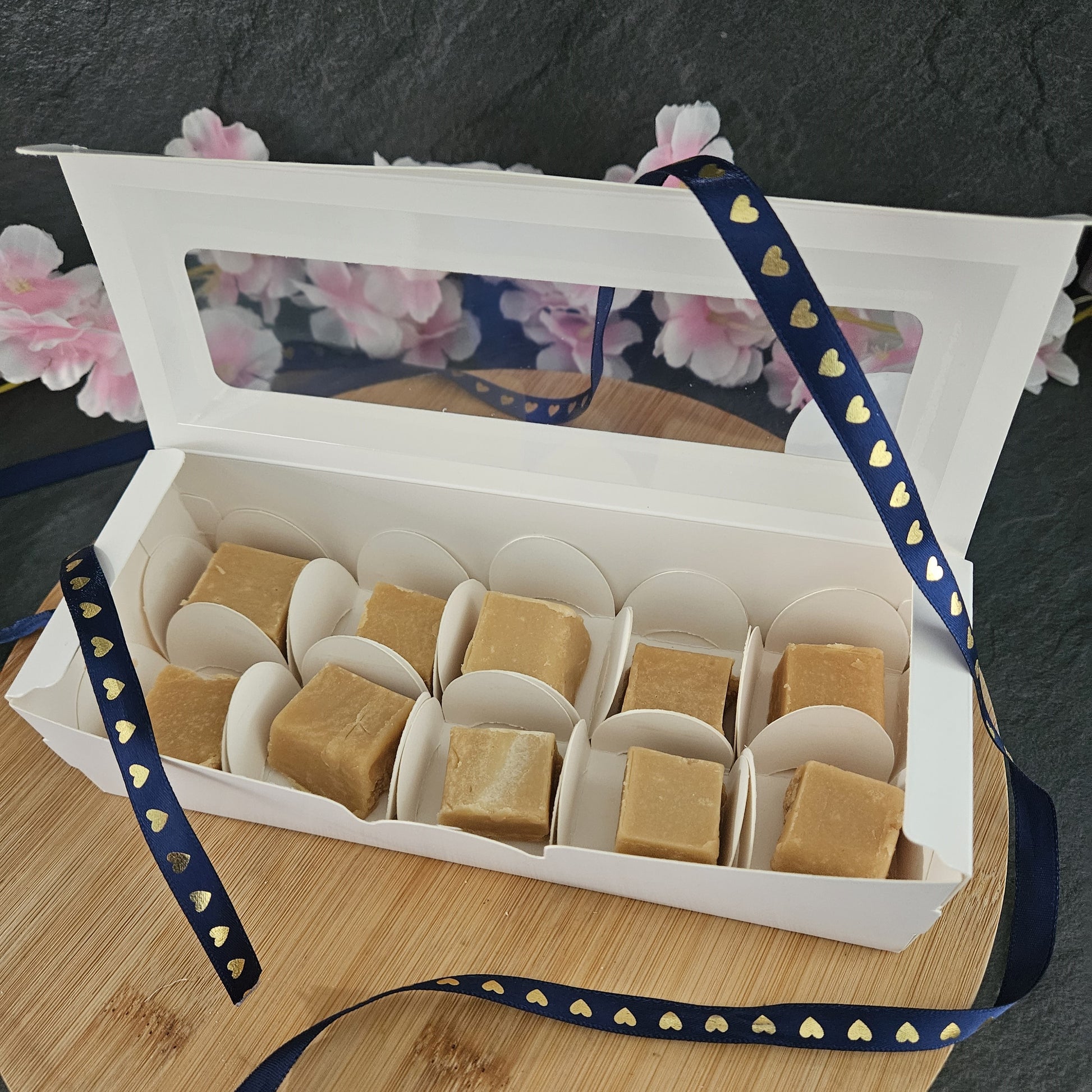 Open white box f fudge with 10 pieces. sat on wooden board with pink flowers in background draped in blue ribbon with gold hearts.