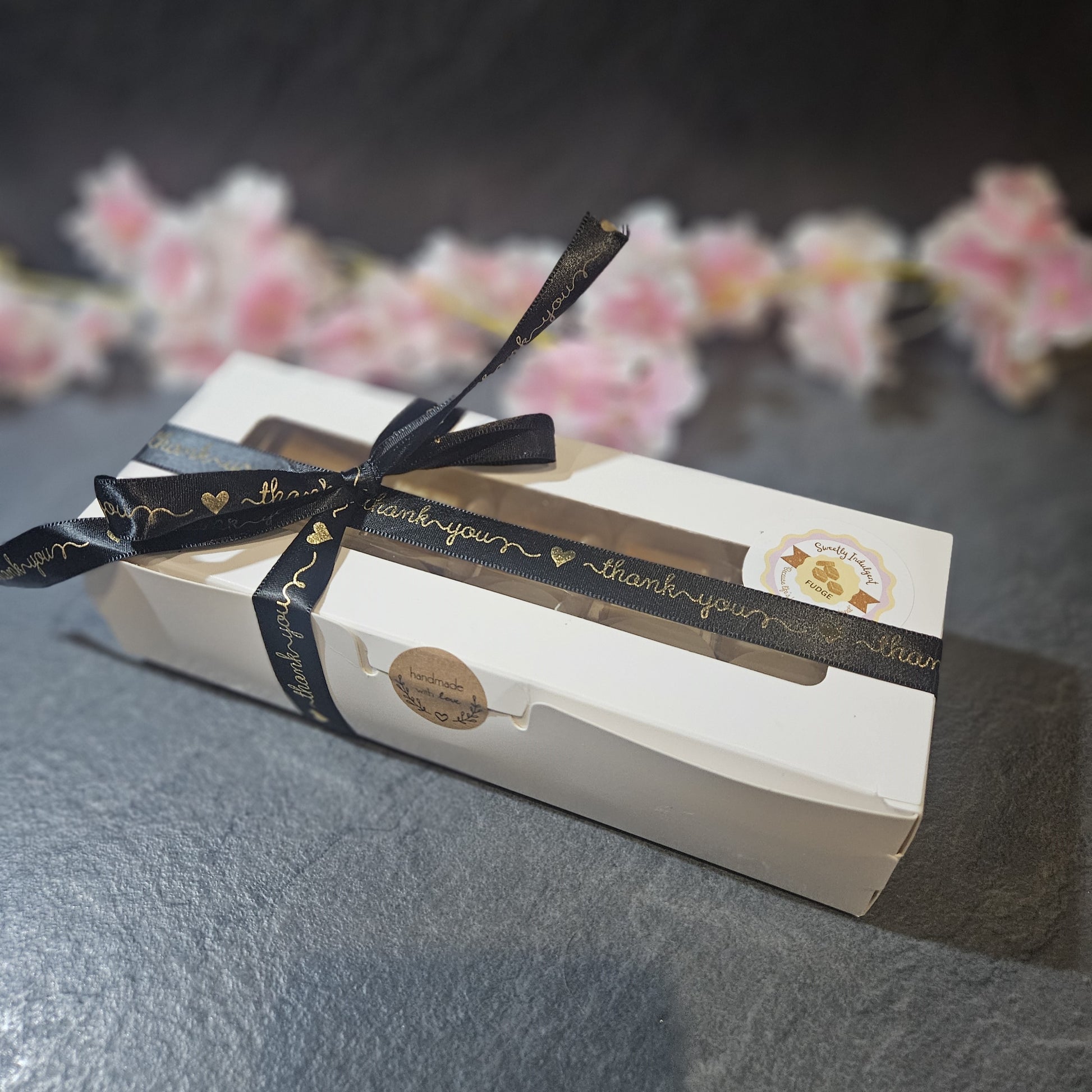 White box with a black ribbon and 'thank you' label on a dark surface with blurred flowers in the background