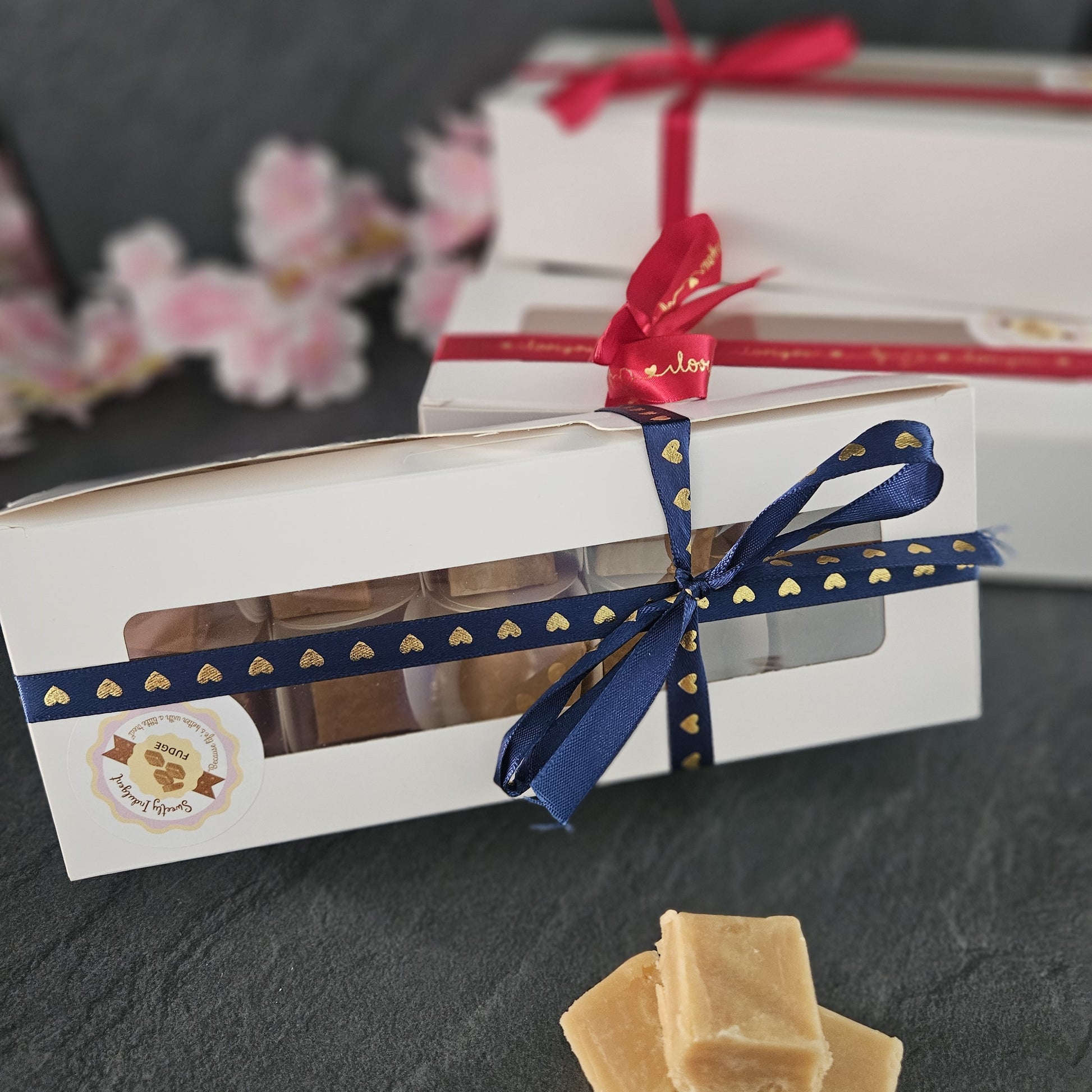 3 gift boxes one with a blue ribbon on a dark surface, red ribbon in background with fudge pieces in the foreground.