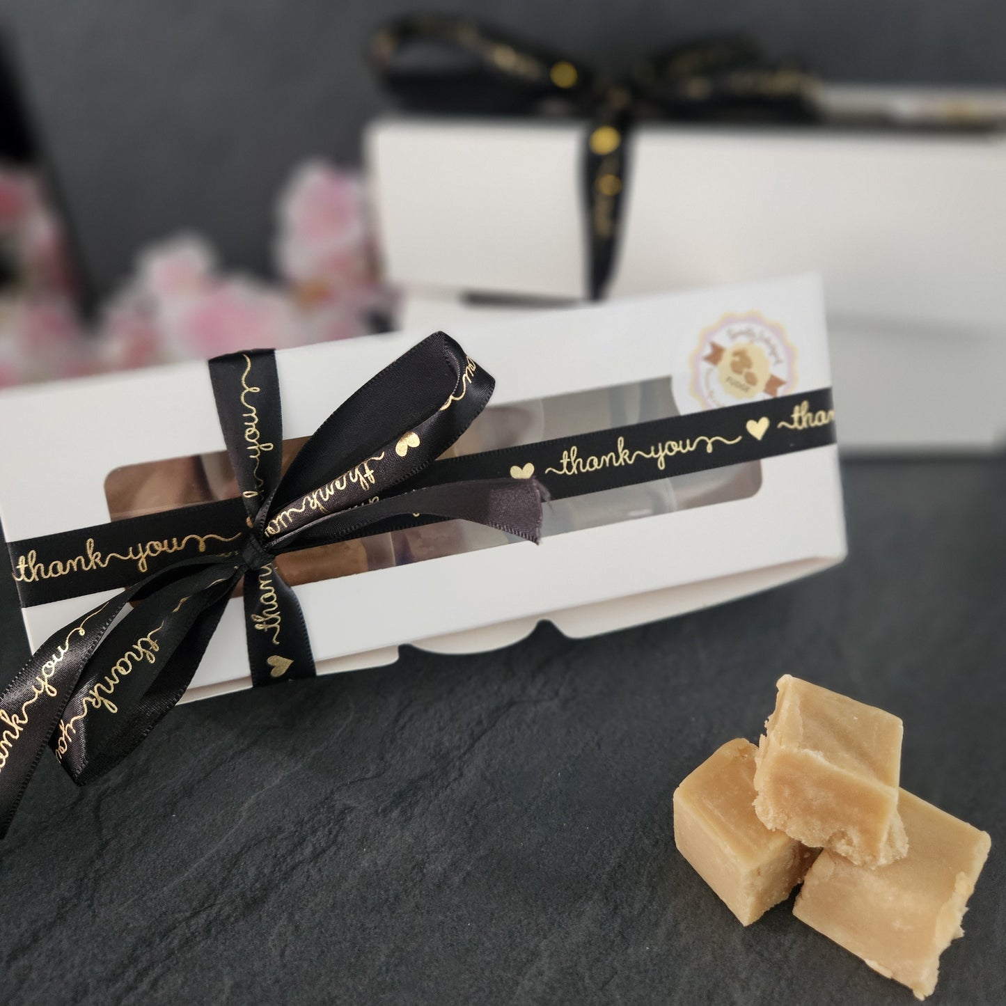 Gift box with a black ribbon and 'thank you' text, accompanied by pieces of fudge on a dark surface.
