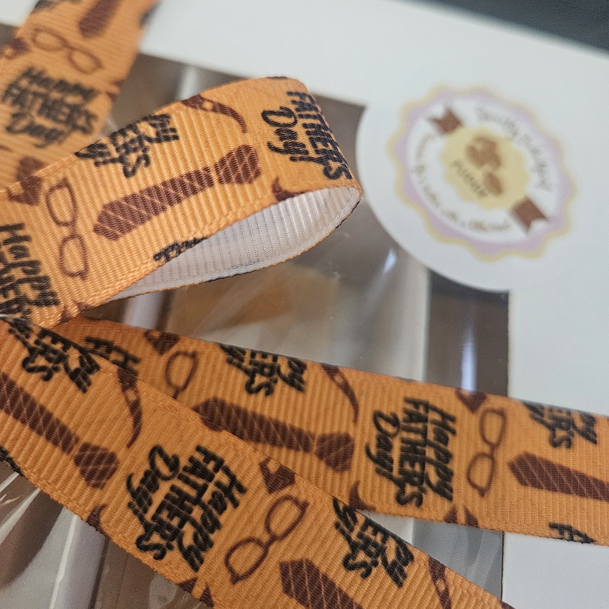 Orange ribbon with 'Happy Father's Day' text on a blurred background with Sweetly Indulgent logo .