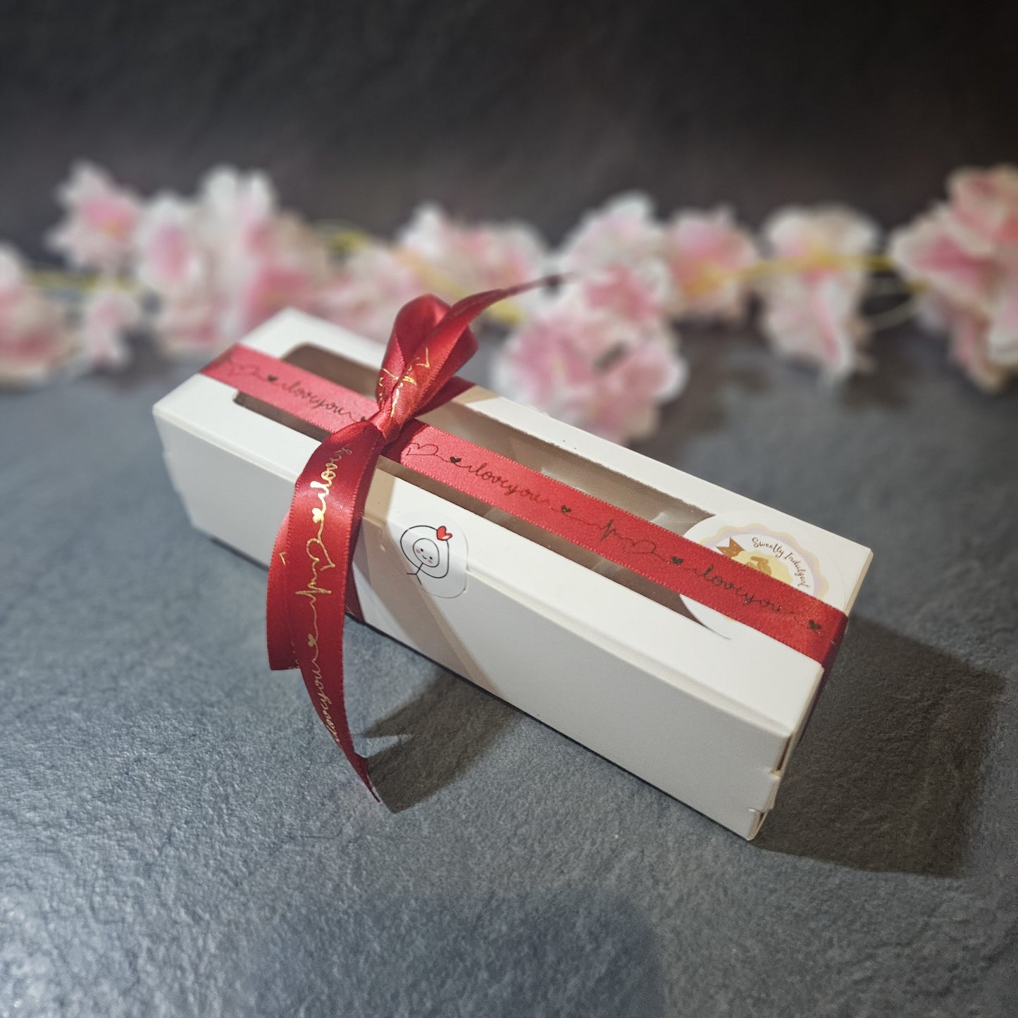 White rectangular box with a red ribbon on a gray surface with blurred flowers in the background