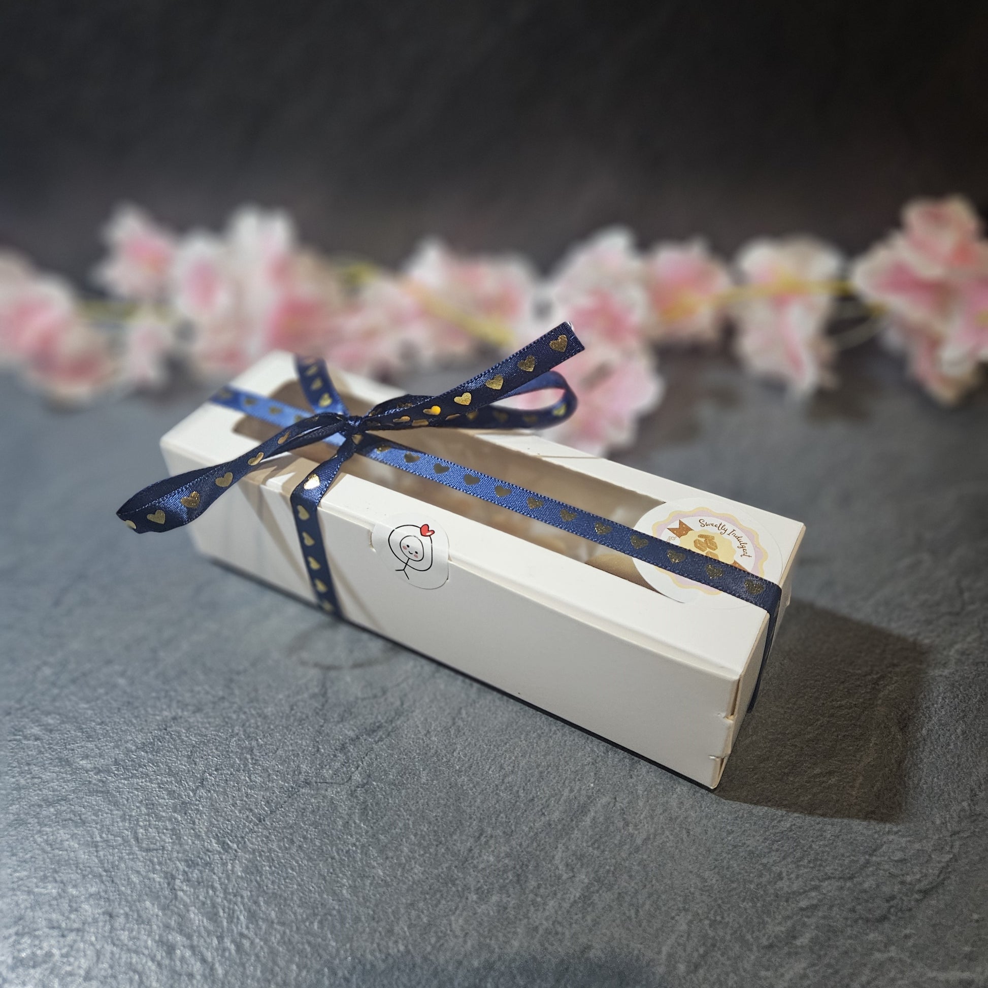 Gift box with a blue ribbon on a dark surface with blurred flowers in the background. sealed with love heart stick man.