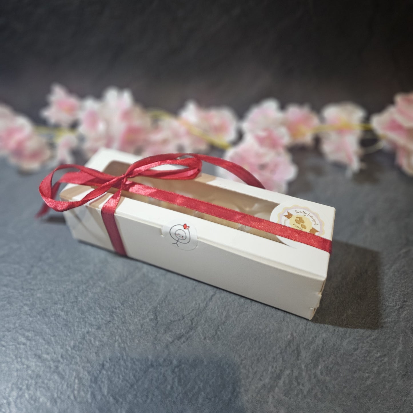 White box with a red ribbon on a dark surface with pink flowers in the background
