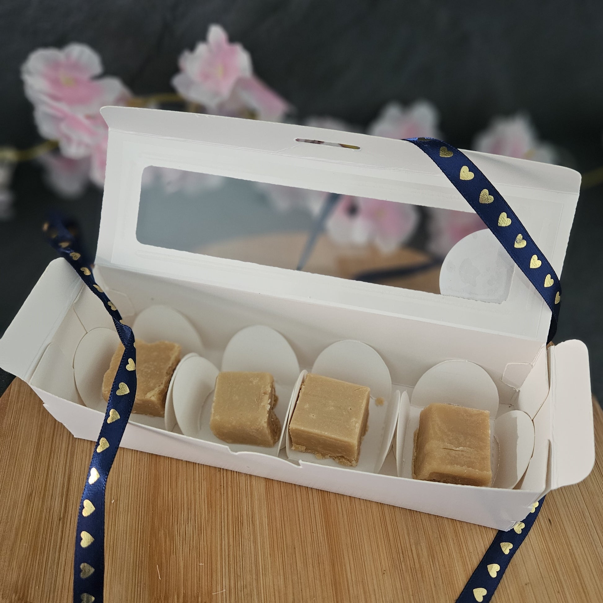 White box with fudge pieces on a wooden surface draped ith blue ribbon.