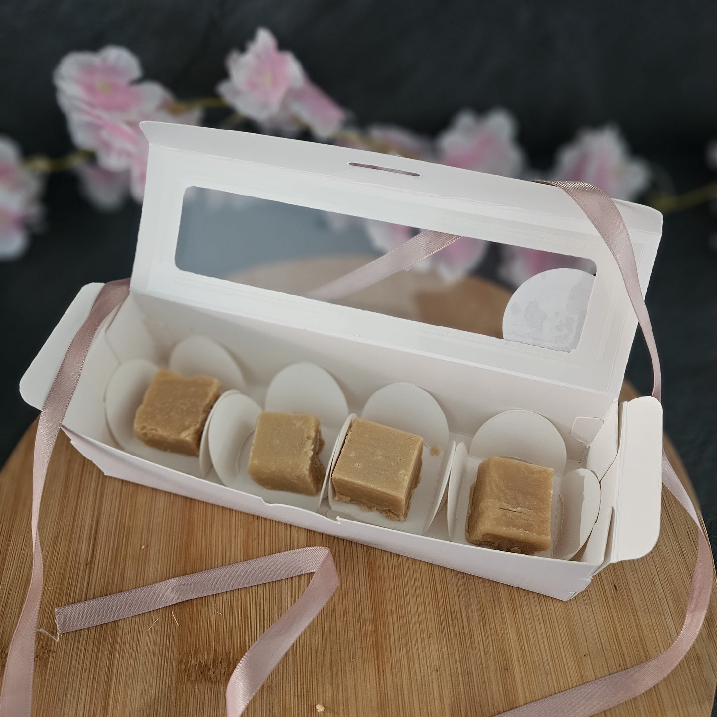 Open white gift box with fudge pieces on a wooden surface with pink ribbons.