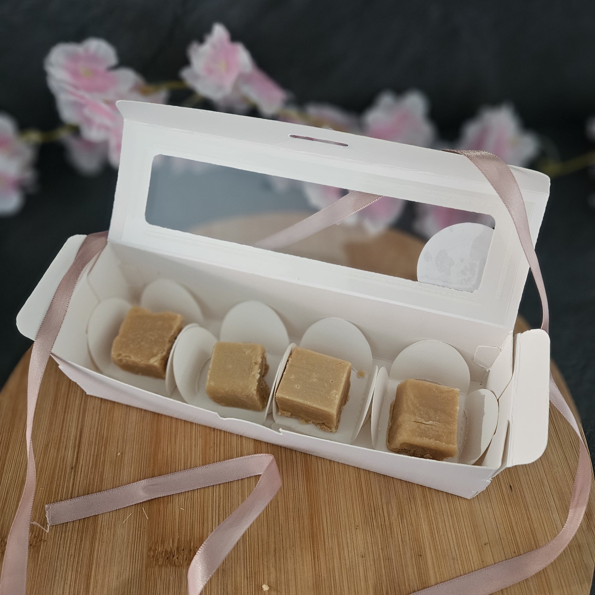 Open white gift box with fudge pieces on a wooden surface with pink ribbons.