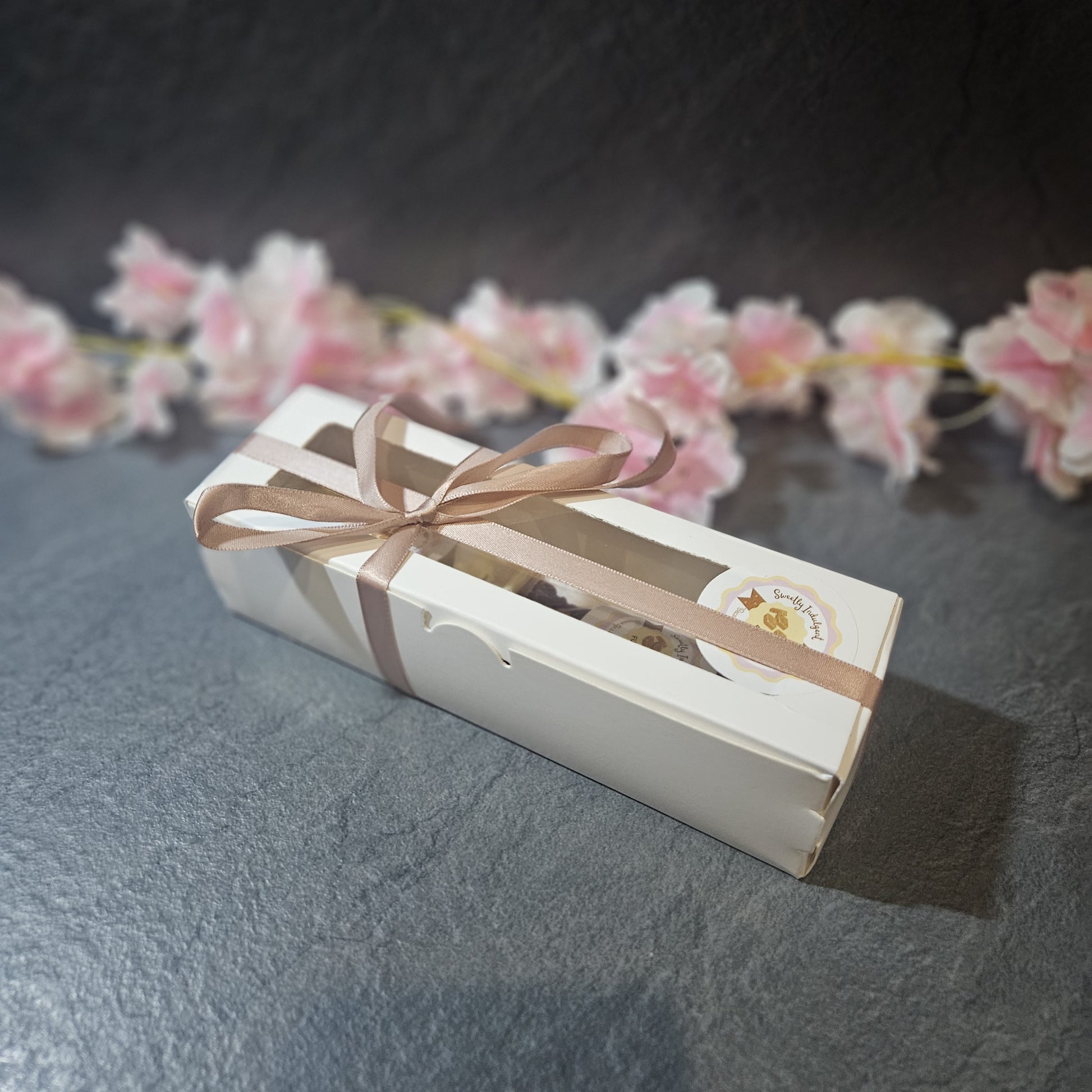 Rectangular gift box with a ribbon on a dark surface with pink flowers in the background.