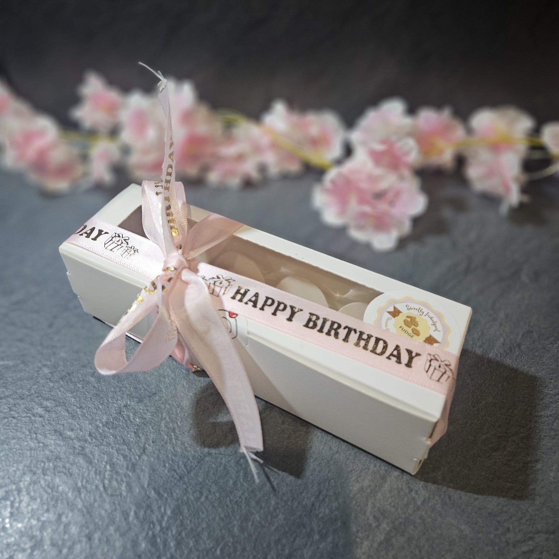 rectangular white box with 'Happy Birthday' text and pink ribbon on a dark surface with floral decorations.