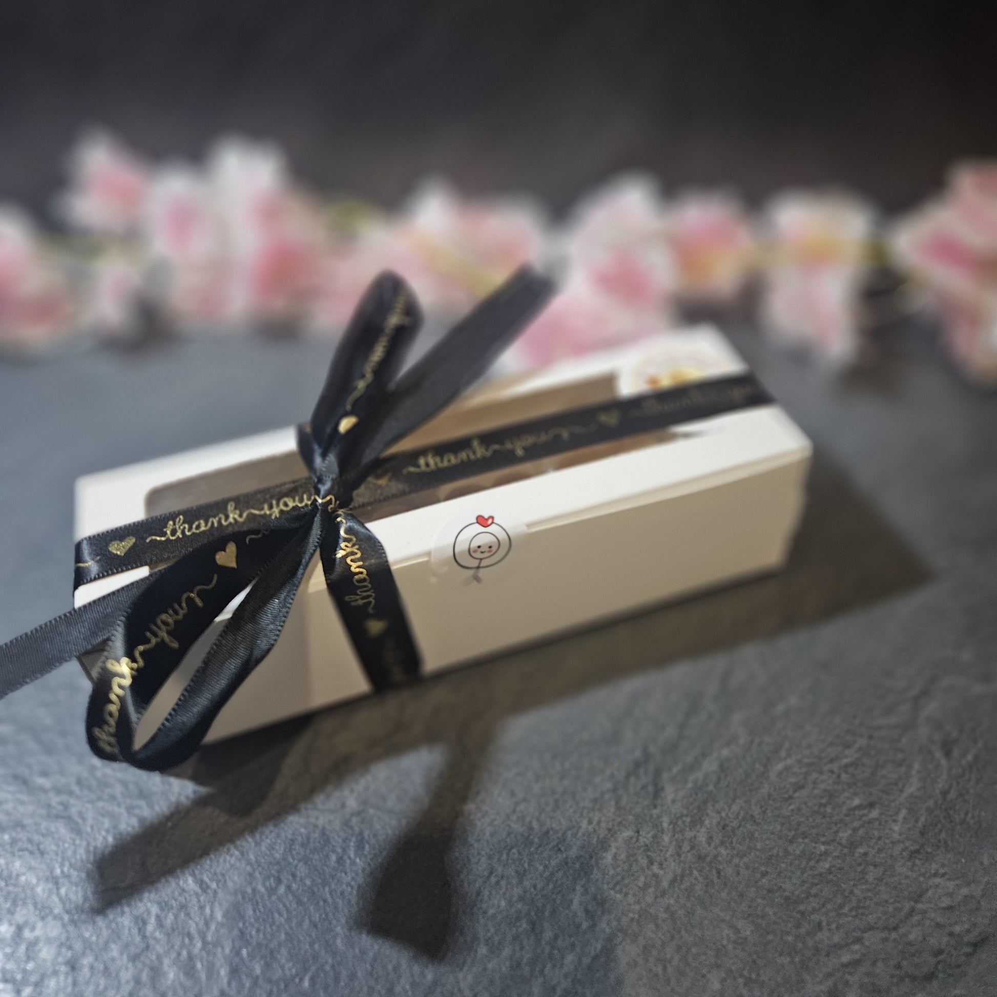 Gift box with a black ribbon and 'thank you' tag on a dark surface with blurred flowers in the background.