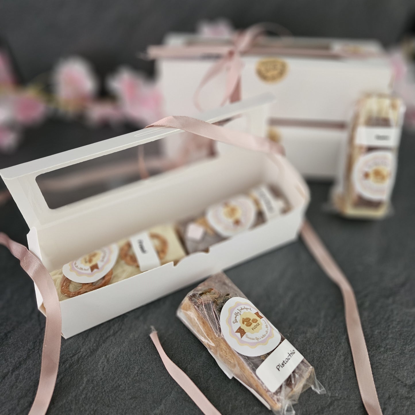 3 white boxes containing 2 fudge topping bars with pink ribbons on a dark surface. fudge bars in foreground.