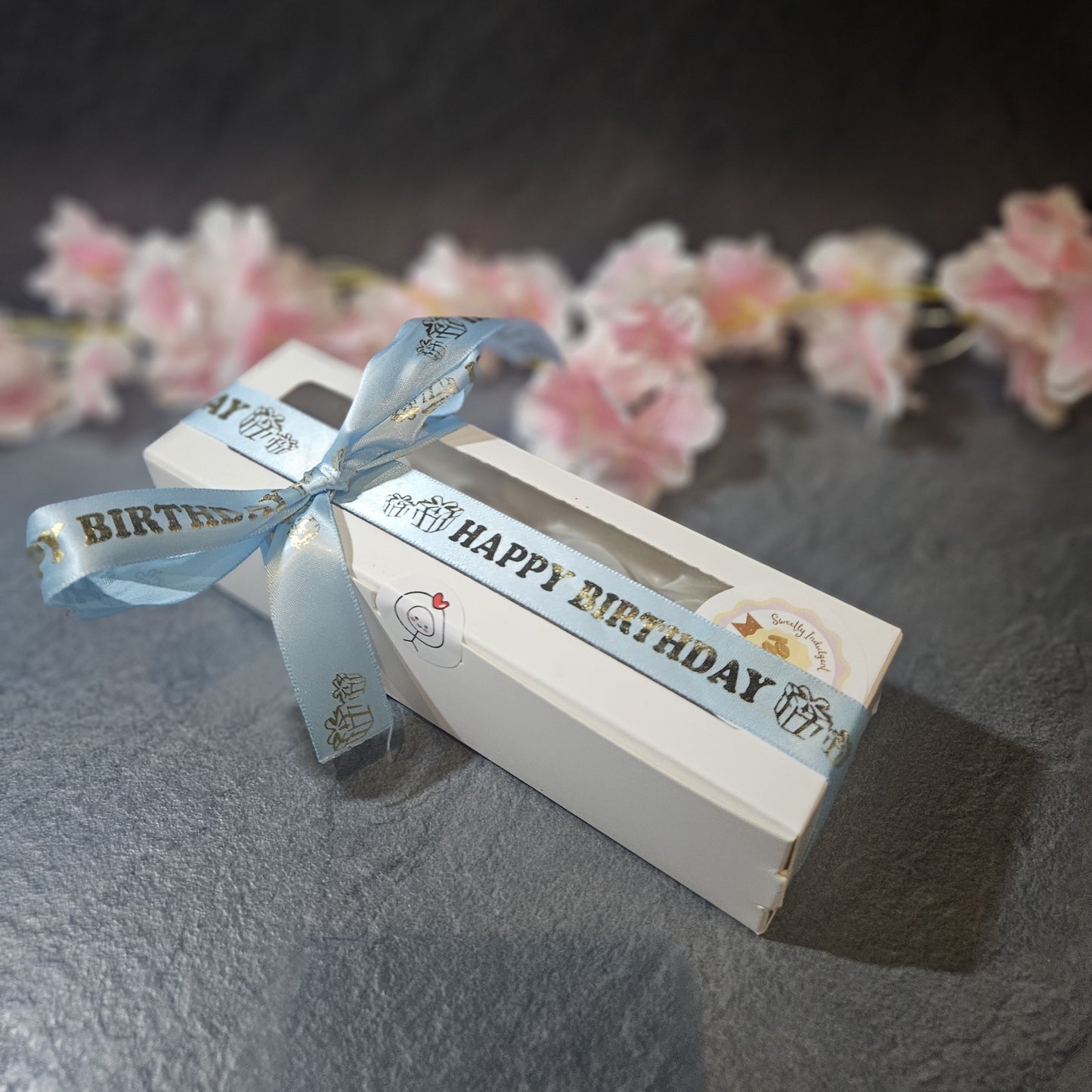 Gift box with Happy Birthday blue ribbon on a dark surface with pink flowers in the background