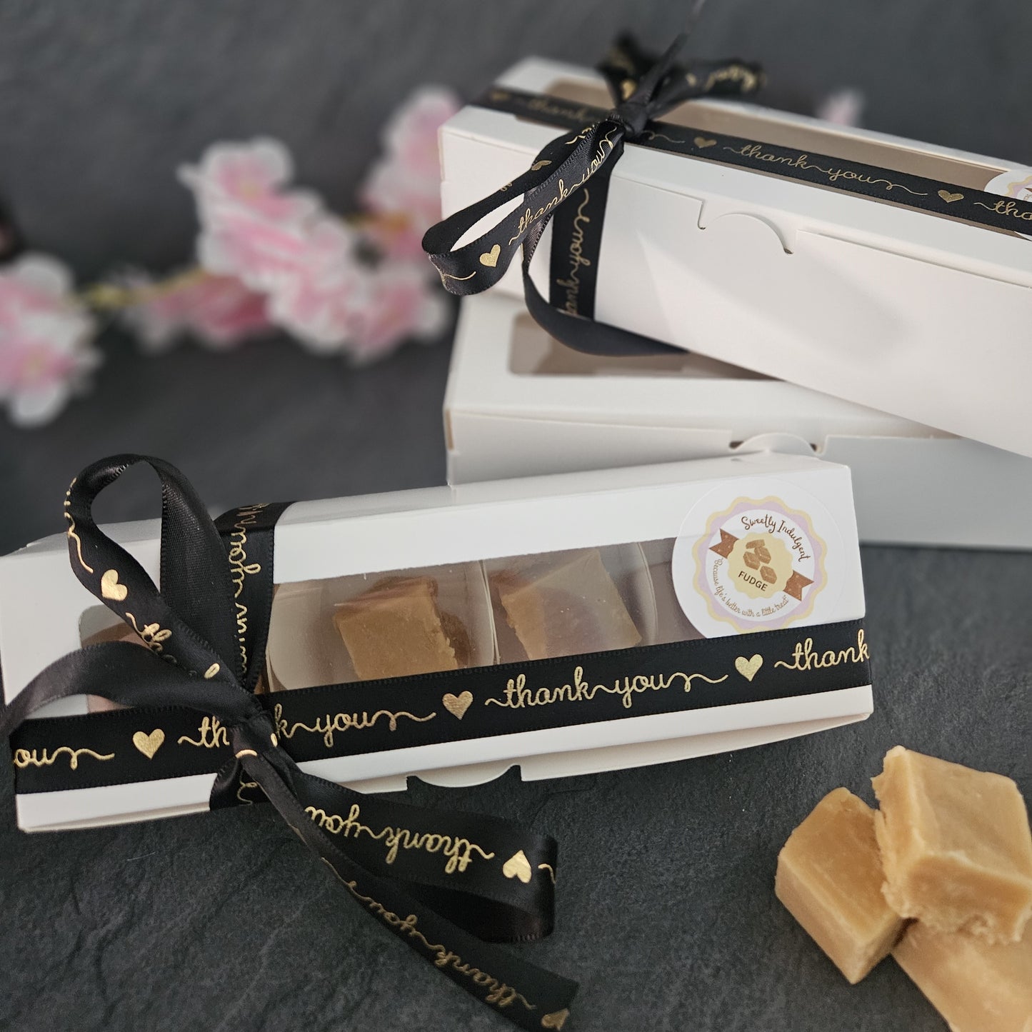 Small box of fudge with a black ribbon and 'thank you' text on a dark surface. Fudge pieces in foreground. 
