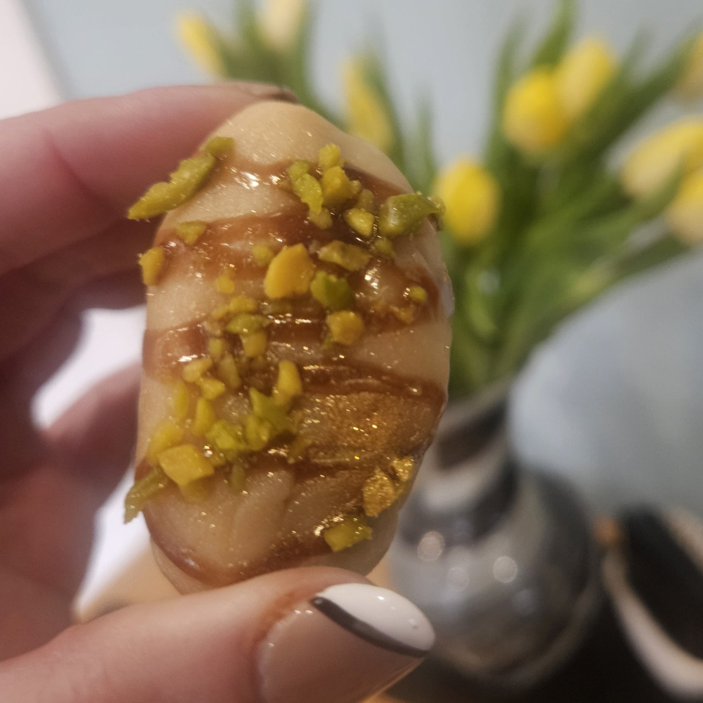Hand holding a fudge dipped date with pistachios and chocolate drizzle in front of a blurred floral background